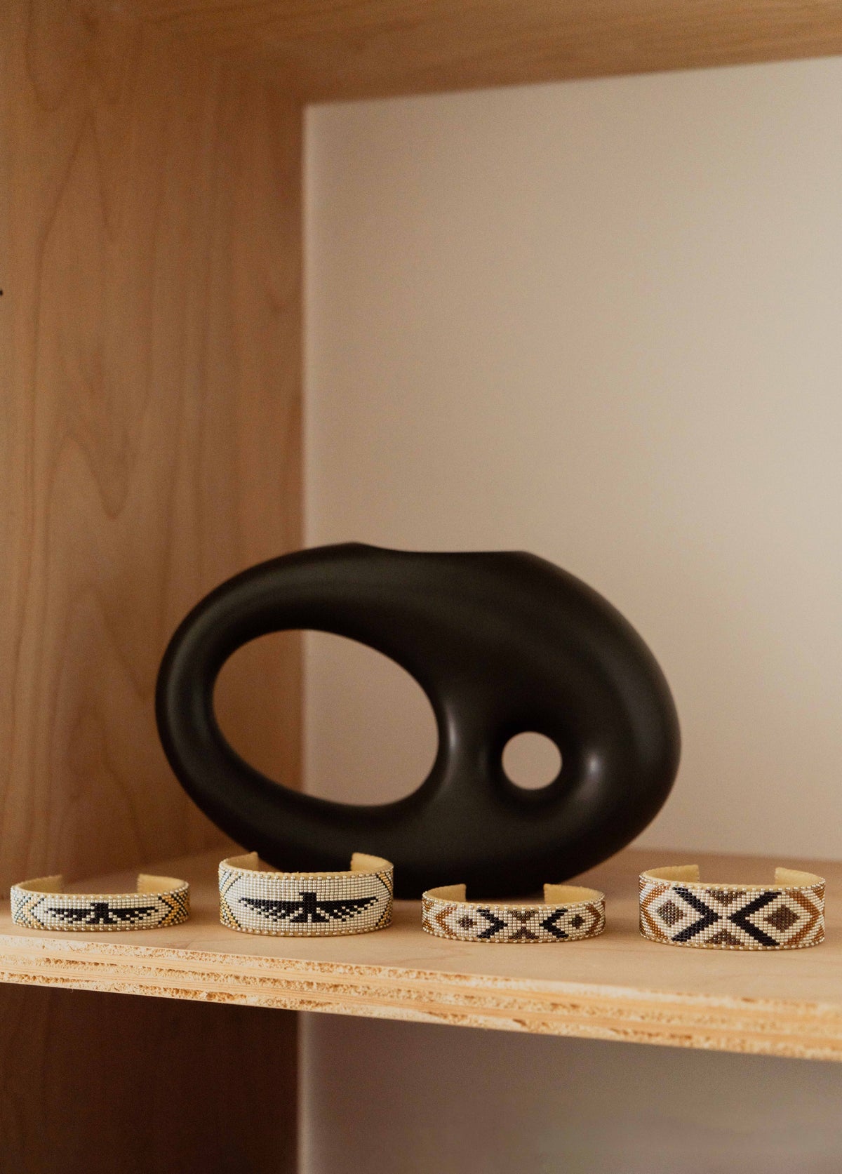 Four Etkie beaded cuffs on a plywood shelf beside a black ceramic vessel
